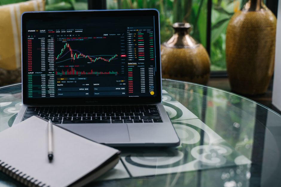 Laptop displaying cryptocurrency stocks and graphs on a glass table with a notepad to illustrate prop firm trading without time limits