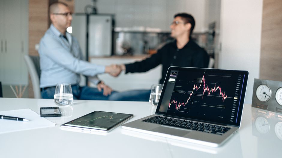 Two businessmen shake hands at a desk with stock market graphs on a laptop to illustrate prop firm affiliate program benefits